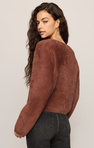 Woman wearing a brown sweater and dark jeans against a plain background