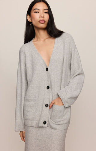 A woman wearing a light gray knit cardigan with a deep V-neck, black buttons, and front patch pockets, paired with a matching gray skirt, against a solid beige background.