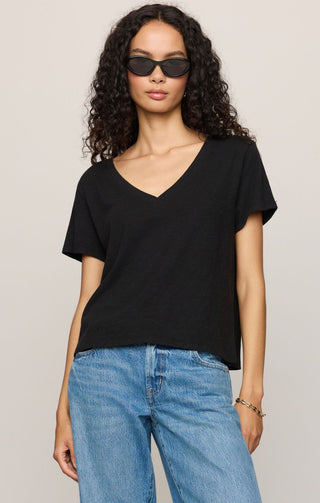 A woman with curly dark hair wearing a black short-sleeve V-neck t-shirt and light blue jeans, standing against a plain light beige studio background.