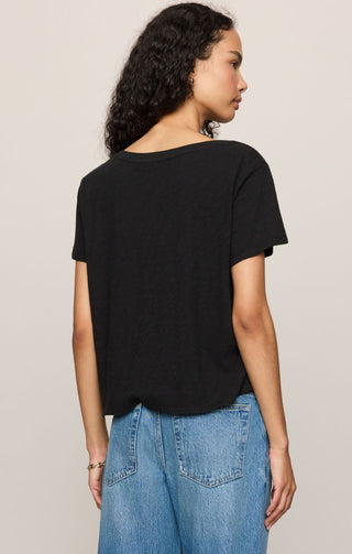 A woman shown from the back wearing a black short-sleeve t-shirt and light blue jeans, standing against a plain light beige studio background.