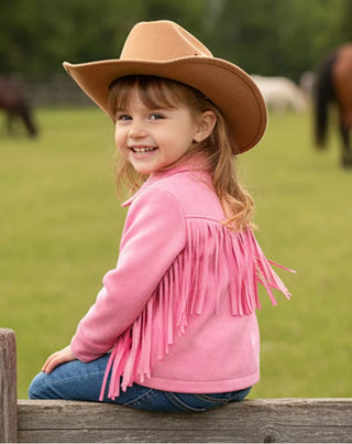 Child wearing a pink fringed jacket and brown cowboy hat sitting on a wooden fence with horses in the background.