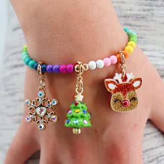 A close-up of a person's hand wearing a beaded bracelet with three dangling holiday charms: a snowflake, a green Christmas tree, and a glittery reindeer head. The bracelet features multicolored beads and gold-tone metal.