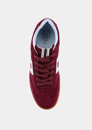A top-down view of a low-top sneaker in burgundy suede with white side stripes, burgundy laces, and a white inner lining, shown on a light gray background.