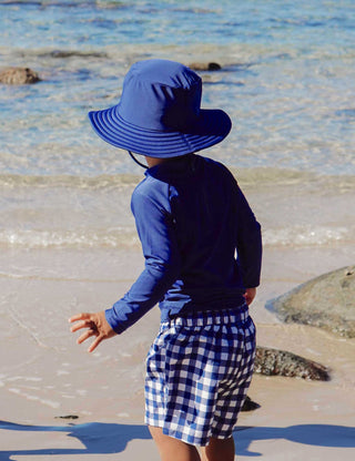 Person wearing a blue sun hat and checkered outfit by a body of water.