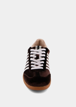 A single low-top sneaker shown from a front-facing perspective against a light gray background. The shoe features a dark brown suede upper with black and white striped side panels, white laces, and a brown gum sole.