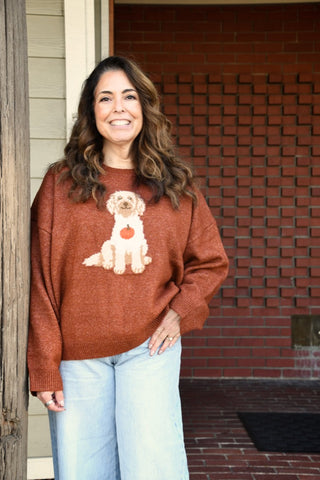 A smiling woman with long brown hair stands on a porch wearing a rust-orange knit sweater with a large white dog graphic on the front, paired with light blue jeans.