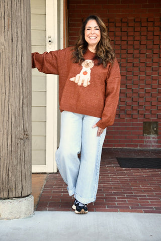 A woman stands on a brick porch wearing a rust-brown knit sweater with a dog graphic on the front, paired with light-wash wide-leg jeans and black sneakers.