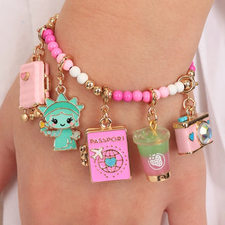 A close-up of a person's wrist wearing a beaded charm bracelet with multiple whimsical charms, including a pink suitcase, a teal cartoon character, a pink passport, a green drink cup, and a pink camera.