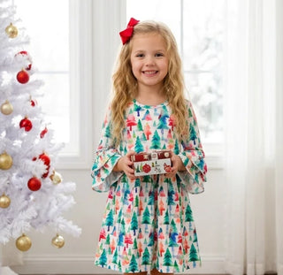 A young girl with a red bow in her hair stands in a bright room wearing a white dress with a colorful Christmas tree print and holding a wrapped gift. A decorated white Christmas tree is visible on the left.