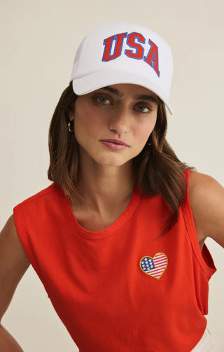 Woman wearing a white cap with 'USA' and a red tank top with a heart design on a beige background