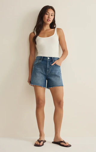 Woman wearing a white tank top and blue denim shorts against a beige background