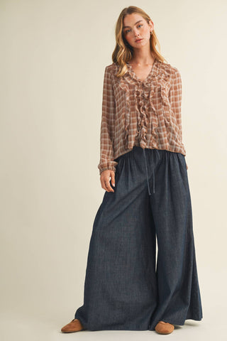 A woman modeling a long-sleeve ruffled blouse in a brown and beige plaid pattern, paired with wide-leg dark blue pants and brown shoes, against a plain beige studio background.
