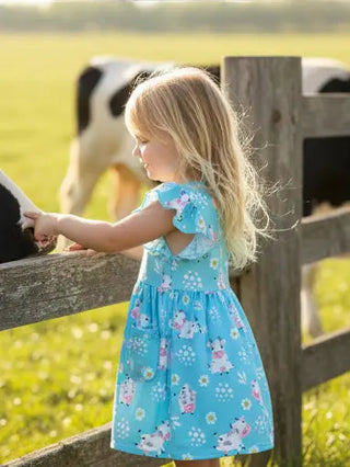 Child in a blue dress standing near a wooden fence with cows in the background