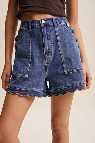 Blue denim shorts with scalloped hem and gold buttons on a neutral background