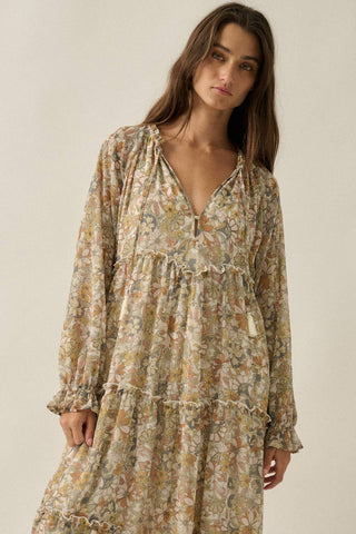 A woman wearing a long-sleeved, tiered bohemian-style maxi dress with a dense floral print in beige, green, and brown tones, set against a plain light beige background.