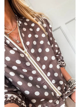 A close-up of a woman wearing a brown and white polka dot zip-up jacket with leopard-print cuffs, layered with silver necklaces and bracelets.