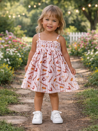 Child in a patterned dress standing on a garden path with flowers and a white picket fence in the background.