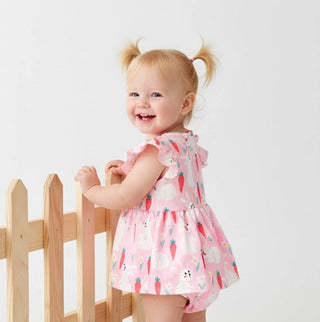 Child wearing a pink dress with a carrot pattern, standing next to a wooden fence on a light gray background