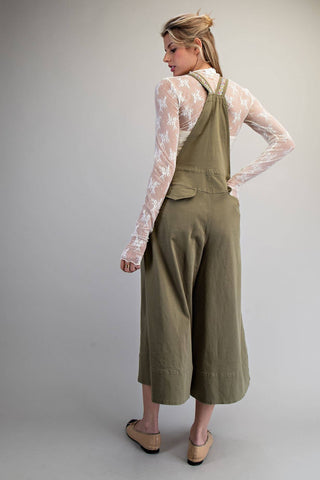 A woman modeling olive green wide-leg overall pants with a sheer white floral long-sleeve top, shown from a back-three-quarter angle against a neutral gray studio background.