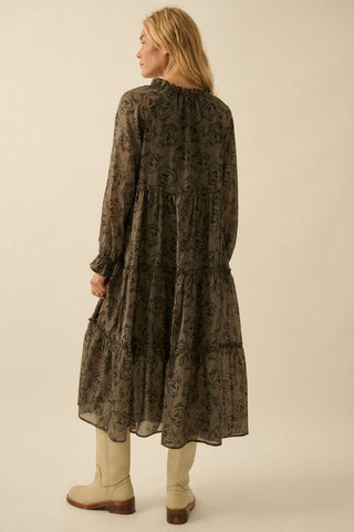 A woman shown from the back wearing a long-sleeve, tiered midi dress with an all-over dark floral lace pattern in olive and black, paired with beige knee-high boots, against a solid light beige studio background.