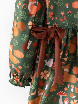 A close-up of a long-sleeve dress with a forest-themed print featuring squirrels, foxes, deer, mushrooms, and leaves on a dark green background, finished with a brown fabric tie belt at the waist.