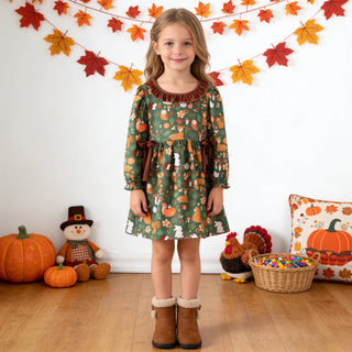 A young girl stands in a decorated room wearing a green long-sleeve dress with a pumpkin and autumn pattern, paired with brown boots. The background features a white wall with colorful fall leaf garlands, pumpkins, a snowman, and a pumpkin-patterned pillow.