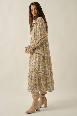 A woman modeling a long-sleeved, tiered midi dress with a dense floral print in beige, yellow, and green tones, paired with beige heeled ankle boots against a solid light beige studio background.