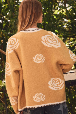 A woman viewed from behind wearing a camel-colored knit sweater with a cream rose pattern, set against a background of green foliage.