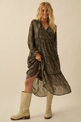 A woman modeling a long-sleeve, tiered midi dress with a dark olive green and black floral pattern, paired with beige boots, against a solid beige studio background.