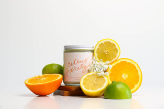 Candle labeled 'Citrus Spritz' surrounded by sliced oranges, lemons, and limes on a white background