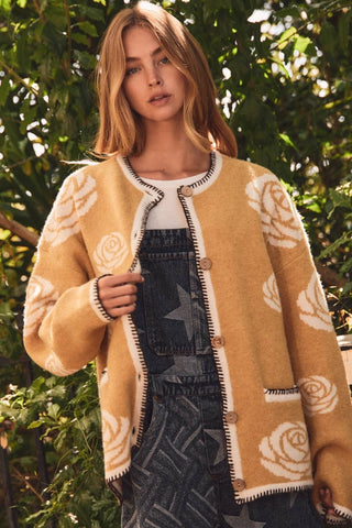 A woman wearing a mustard yellow fuzzy cardigan with cream rose patterns and dark denim overalls, standing outdoors in front of green foliage.