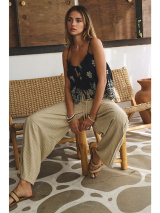 Woman sitting on a wooden chair wearing a floral top and beige pants in a room with woven furniture and a patterned floor.