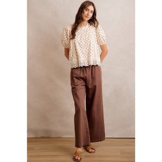 Woman wearing a white blouse with a floral pattern and brown pants against a beige wall.