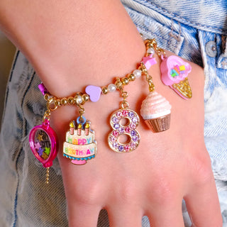 A close-up of a person's hand and wrist wearing a gold charm bracelet with multiple colorful birthday-themed charms, including a pink heart, a birthday cake, an 8 with rhinestones, a cupcake, and a pink ice cream cone, styled with light blue denim jeans.