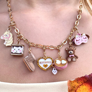 A close-up of a gold-tone charm necklace worn on a person's neck. The necklace features multiple cute charms including a horse, a hamster, a marshmallow, a boot, a heart with 'NOVEMBER' inside, a cupcake, a teddy bear, and a small white heart.