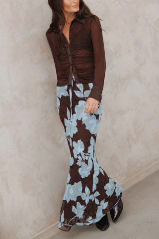 A woman models a two-piece outfit featuring a dark brown ruched long-sleeve top and a matching dark brown maxi skirt with a light blue floral pattern, set against a textured beige wall.