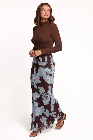 A woman modeling a brown mock-neck long-sleeve knit top paired with a brown maxi skirt featuring a large light gray floral print, against a plain white studio background.