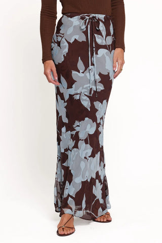 A woman wearing a long brown maxi skirt with a large light gray floral print and a drawstring waist, paired with a brown long-sleeve top and brown strappy sandals, against a plain white background.