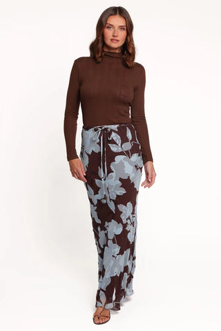 A woman modeling a brown mock-neck long-sleeve top paired with brown and light gray floral print pants, standing against a plain white studio background.