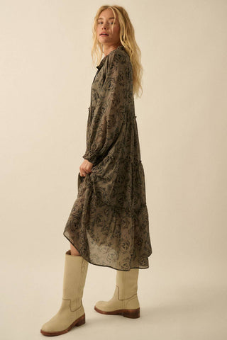 A woman modeling a long-sleeved, midi-length sheer tunic dress with an abstract dark brown and black floral pattern, paired with beige mid-calf boots, against a solid light beige studio background.