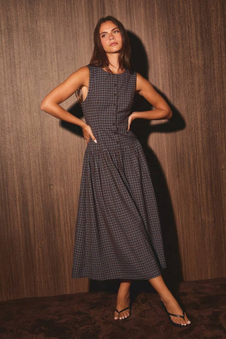 A woman modeling a sleeveless, knee-length plaid dress with a buttoned bodice and flared skirt, standing against a dark wood-paneled wall.