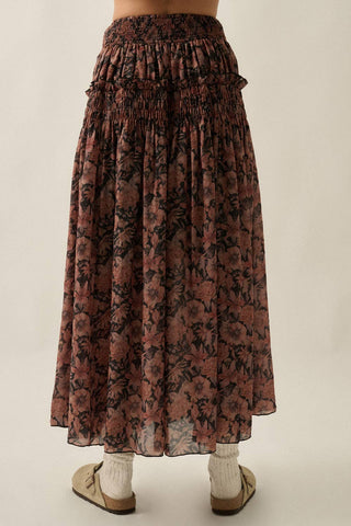 A long, gathered midi skirt with a dark floral print in dusty rose and black, shown on a model from the back against a plain beige background.