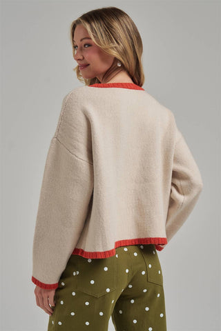A woman shown from the back wearing a cropped knit sweater in a warm cream color with red trim, paired with olive green pants with white polka dots, against a solid gray studio background.