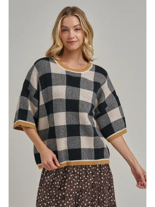 A woman modeling a black and off-white checkered knit sweater with mustard yellow trim, paired with a brown polka dot skirt, against a solid gray studio background.