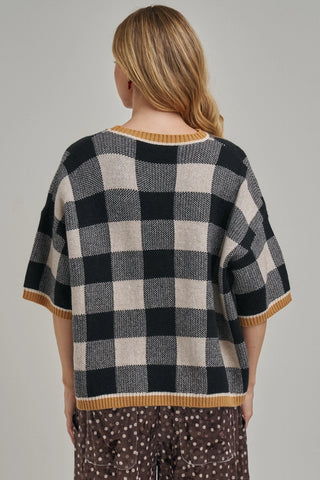 A woman viewed from the back wearing a black and cream buffalo check knit sweater with mustard yellow ribbed trim, paired with brown patterned pants, against a solid light gray background.