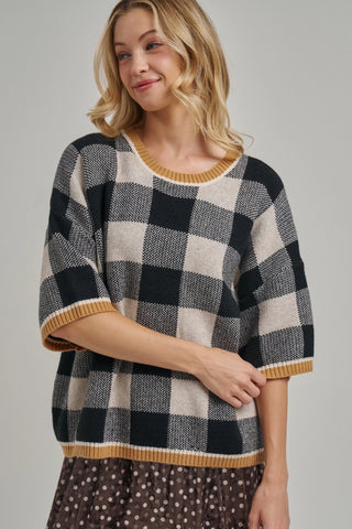 A woman wearing a black and cream buffalo check knit sweater with mustard yellow ribbed trim on the crew neck, sleeve cuffs, and hem, paired with a brown polka dot skirt, against a solid light gray background.
