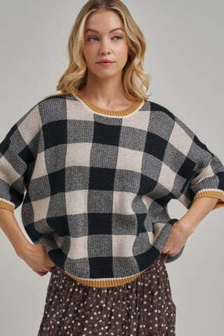 A woman wearing a black and beige buffalo check knit sweater with mustard yellow trim, paired with a brown polka dot skirt, against a solid gray studio background.