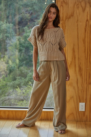 Woman wearing a beige knit top and pants standing in a room with wooden walls and a window showing greenery outside.