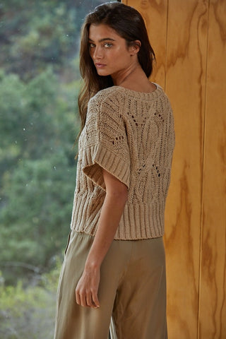 Woman wearing a beige knitted top and pants standing against a wooden wall with greenery in the background