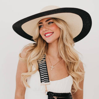 Woman wearing a wide-brimmed hat with a black and white strap over her shoulder on a plain background